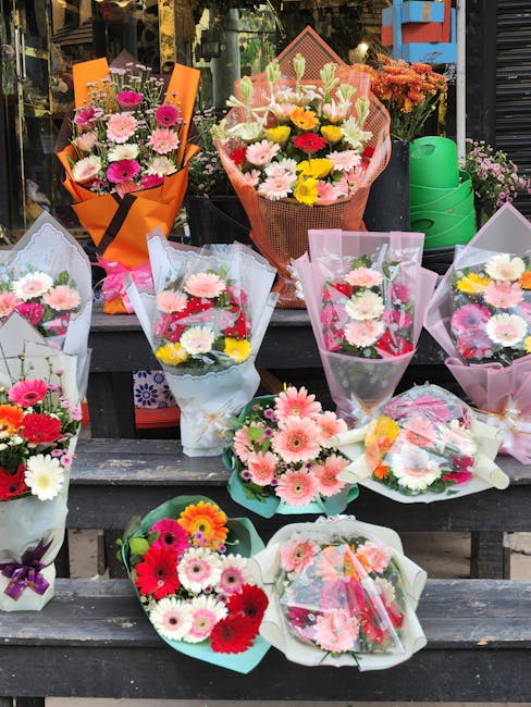 A display of fresh floral arrangements and bouquets placed on wooden steps outside a flower shop. The arrangements feature a variety of flowers including pink gerbera daisies, white chrysanthemums, re
