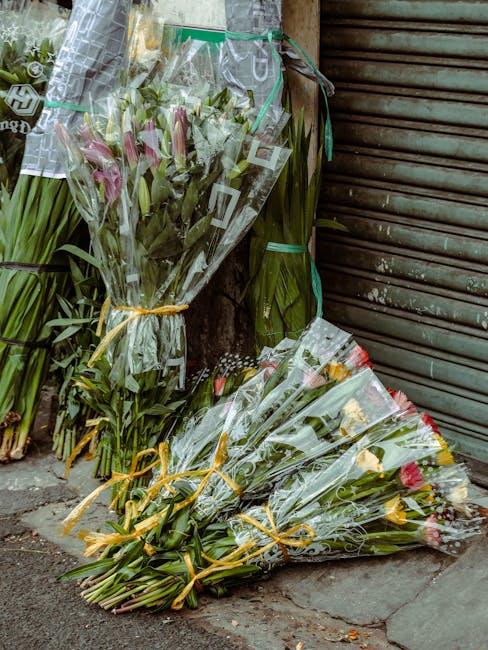 A collection of fresh flower bouquets resting on a sidewalk in front of a closed metal shutter. The arrangements feature pink lilies, yellow tulips, and other mixed blooms, all wrapped in clear cellop