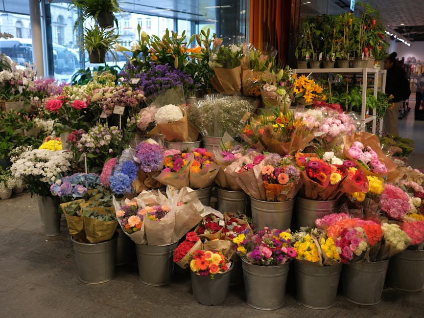A vibrant display of floral arrangements and bouquets inside a florist shop, featuring a variety of fresh flowers including pink roses, purple statice, white daisies, yellow sunflowers, and multicolor