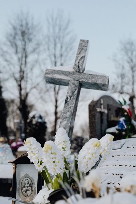 A floral tribute featuring fresh white flowers, likely chrysanthemums or lilies, arranged in a bouquet with green stems visible, placed at the base of a grey, stone cross grave marker. The flowers app