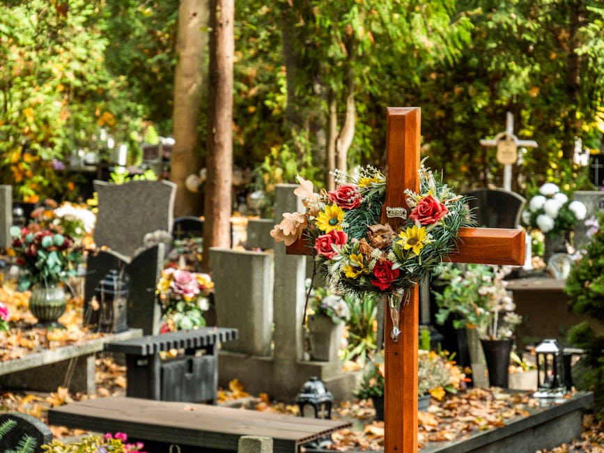 A wooden cross decorated with an arrangement of fresh flowers, including pink roses, yellow daisies, pale peach blooms, and green foliage, is positioned in a cemetery surrounded by various headstones 