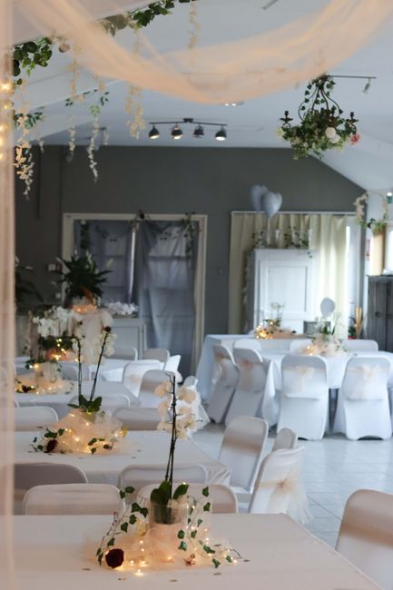 A decorated event space featuring elegantly arranged floral centerpieces on white tablecloths, with tall slender vases holding white orchids and dark red roses, surrounded by fairy lights. The ceiling