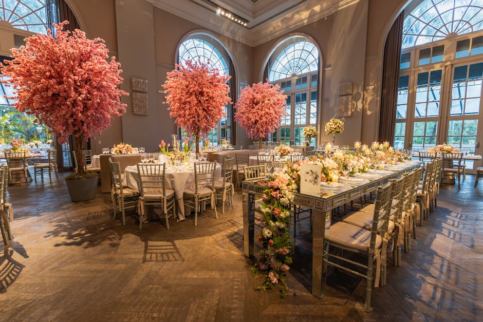 An elegantly decorated banquet hall featuring a long central table adorned with a floral arrangement of pastel pink and white flowers, including roses and peonies, complemented by numerous candles in 