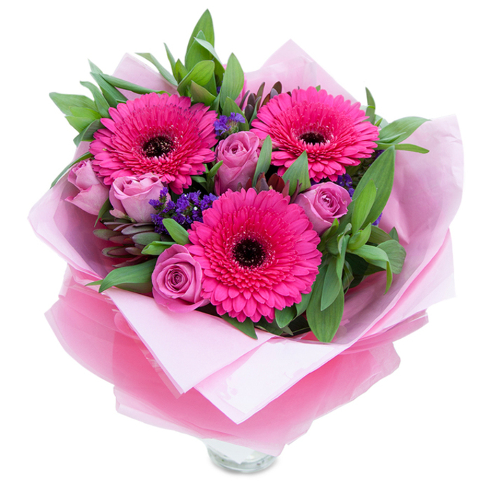 A graceful bouquet captured on a plain white background, where playful fuchsia and gentle lavender tones meet in a composition that feels both garden-fresh and thoughtfully crafted for the Charing Cross neighbourhood. Three wide-faced Gerbera daisies in a confident fuchsia pink take centre stage, their concentric petals spreading like small, vivid suns around deep dark centres that add a poised contrast. Interwoven among these are softly blushing roses in dusty rose to lavender-pink shades, their petals still tightly wound in the early stages of bloom and offering a smooth, velvety texture that invites close appreciation. Small clusters of purple-blue statice appear as tiny islands of colour, bringing an airy, meadow-like quality and a textural counterpoint to the smoother roses and gerberas. Generous green foliage-broad, glossy leaves akin to salal or eucalyptus-creates a verdant frame that lifts the palette and gives a fresh, slightly dewy impression.
The bouquet is wrapped with a florist's attention to layering: a soft, matte pale pink tissue cradles the stems, and over it a sheer, slightly crinkled fuchsia-pink cellophane adds a whisper of shimmer and cohesion to the pink theme. The visible top rim of a clear glass vase at the base signals this arrangement is prepared for immediate display and enjoyment, its stems ready to take up water and renew the blooms. In soft daylight the petals read as tactile-velvet, satin and crisp paper-and the scent suggests a gentle floral mix, roses leading with a delicate perfume while gerberas contribute a light, green sweetness.
This bouquet would sit beautifully on a café table near Villiers Street or as a thoughtful gift delivered to a Flat near Northumberland Avenue, perfect for birthdays, anniversaries, Mother's Day or simply to say 'you matter.' It's a piece that marries playful colour with gracious craftsmanship.